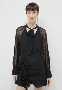 Frau trägt eine schwarze Bluse mit Spitzenärmeln und einem Halsband mit Schleife, kombiniert mit einem gerafften schwarzen Minirock mit Rüschendetails.