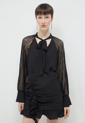Donna che indossa una blusa nera con maniche in pizzo e un colletto a fiocco, abbinata a una mini gonna nera arricciata con dettagli a ruffle.