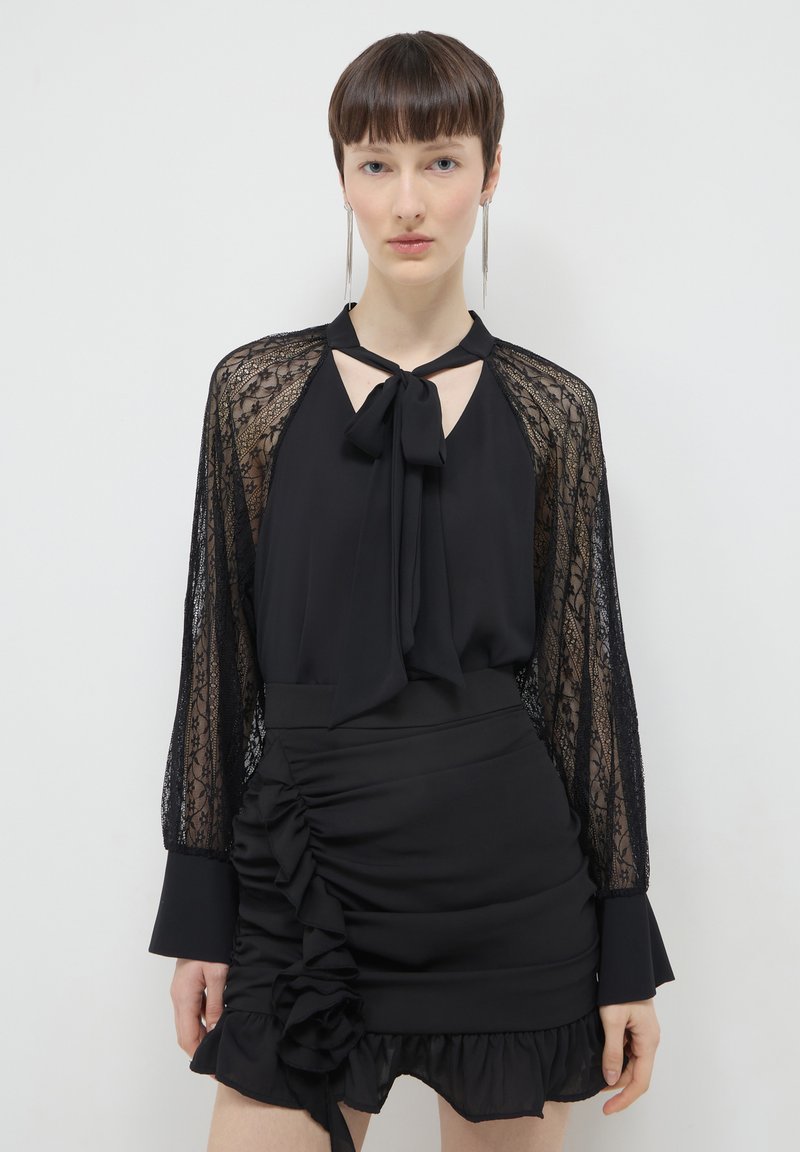 Frau trägt eine schwarze Bluse mit Spitzenärmeln und einem Halsband mit Schleife, kombiniert mit einem gerafften schwarzen Minirock mit Rüschendetails.