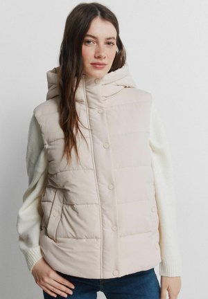 Kvinde med langt brunt hår iført en lys beige quiltet hættetrøje over en hvid sweater og blå jeans, stående med hånden på hoften.