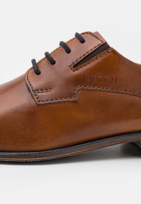 Chaussure en cuir marron à finition lisse, lacets noirs, logo embossé et détails de couture. Présente un bout arrondi et un petit talon.
