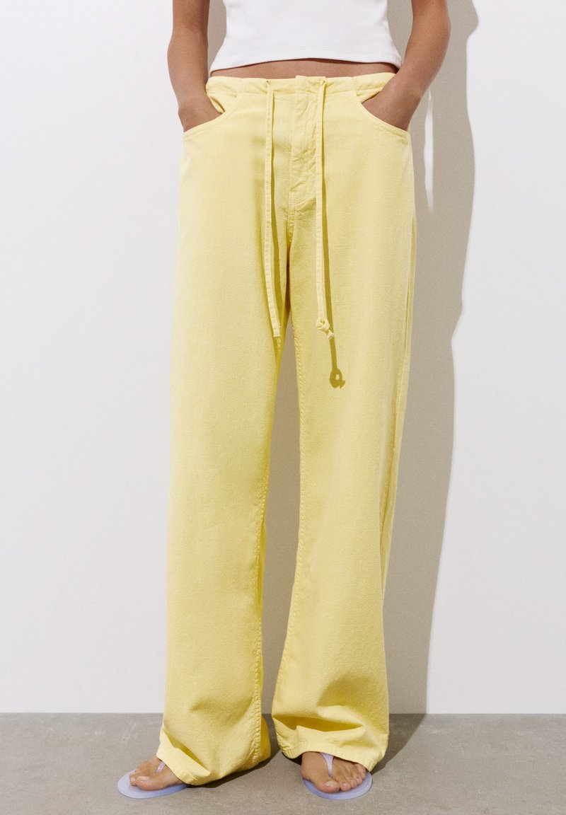Personne portant un pantalon ample jaune à cordon et un top court blanc, debout les mains dans les poches et pieds nus sur un sol gris.