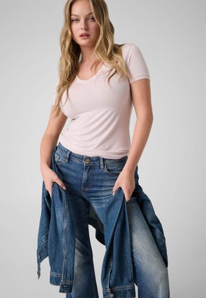 Femme blonde portant un T-shirt moulant rose clair et un jean bleu, tenant une veste en jean devant elle sur un fond uni.