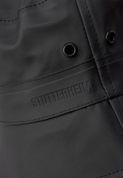 Impermeabile nero con una texture liscia, dotato di cuciture rinforzate, occhielli in metallo e logo "STUTTERHEIM" in rilievo.
