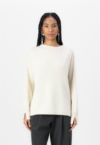 Pull à côtes crème avec une coupe décontractée, un décolleté large et des fentes sur les côtés. Associé à un pantalon gris anthracite pour un look contrasté.