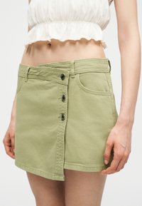 Femme portant une mini-jupe boutonnée asymétrique vert clair et un haut court blanc, dévoilant une partie du ventre et les mains.