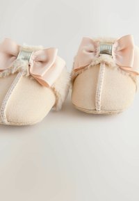 Des chaussons beige clair avec une texture douce, ornés d'un bord duveteux et d'un nœud rayé rose sur chaque chaussure. Détails de marque métallique inclus.