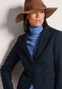 Marineblauwe op maat gemaakte blazer met twee knopen, gedragen over een lichtblauwe coltrui. Een breedgerande bruine hoed voegt een accent toe en toont een gestroomlijnd ontwerp.