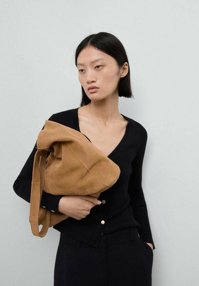 Brun mockaskinn axelväska med en slapp design, hålls av en person som bär en svart ribbad V-ringad cardigan och visar en ren, minimalistisk stil.