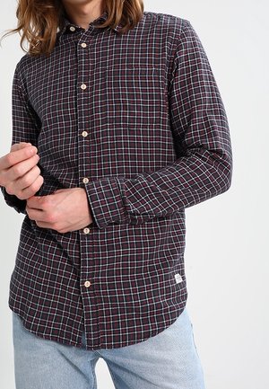 Mann, der ein dunkelrotes und schwarzes kariertes, langärmliges Hemd mit Knopfleiste trägt und die Manschette anpasst, kombiniert mit hellblauen Jeans.