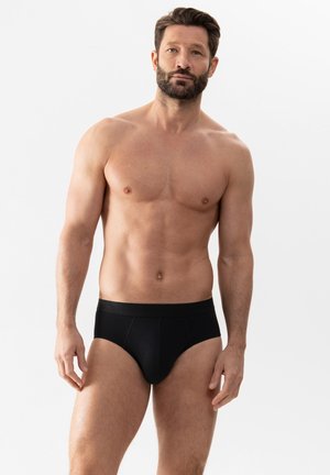 Oberkörperfreier Mann mit kurzen dunklen Haaren und Bart, der schwarze Slips trägt und vor einem schlichten weißen Hintergrund steht.