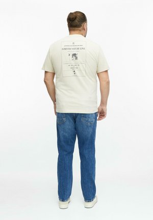 Mężczyzna z kokiem na głowie, ubrany w beżowy t-shirt z tekstem i grafiką o tematyce przyrodniczej na plecach, niebieskie dżinsy i białe trampki, stoi tyłem.
