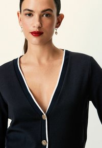Marine cardigan med hvit kant, V-hals og gullknapper. Har et teksturert strikket stof. Nærbillede, der fremhæver halsudskæringen og knapdetaljerne.