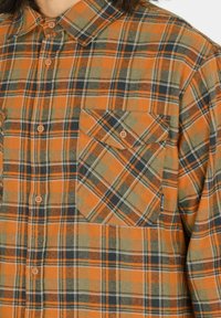 Oranssi ja vihreä ruudullinen flanellipaita, jossa on nappisuljenta, terävä kaulus ja yksi rintatasku nappisuljennalla.