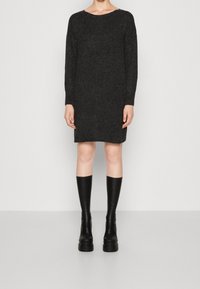 Modelka stojí v černých pletených šatech s dlouhým rukávem, které má zkombinované s černými vysokými kozačkami na podpatku, na bílém pozadí.