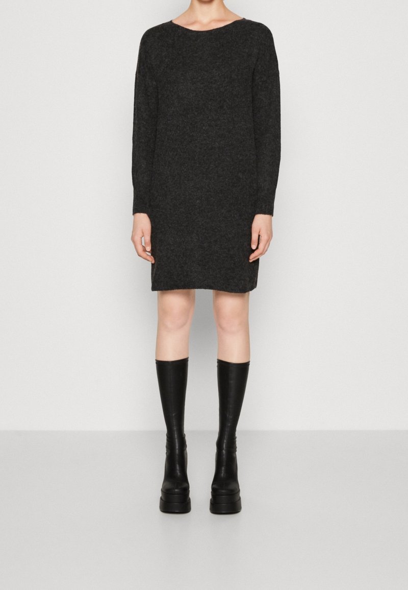 Modelka stojí v černých pletených šatech s dlouhým rukávem, které má zkombinované s černými vysokými kozačkami na podpatku, na bílém pozadí.