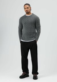 Grijze kabelgebreide trui, losse pasvorm, gedragen over een wit overhemd; zwarte broek met wijde pijpen; zware zwarte sneakers met roze accenten.