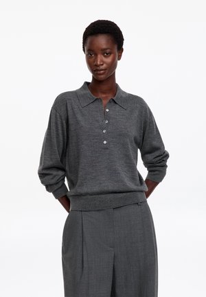 Mujer con el cabello corto, vistiendo una camisa polo gris oscuro con botones y mangas largas y pantalones grises a juego, de pie sobre un fondo blanco.