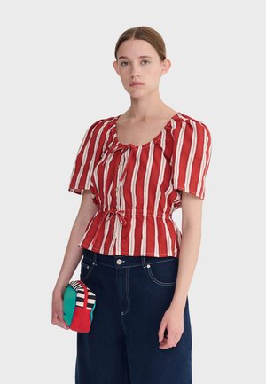 Frau trägt eine kurzärmlige Bluse mit roten und weißen vertikalen Streifen und Bindegürtel, dunkelblaue Jeans, hält ein kleines, mehrfarbiges Täschchen in der linken Hand.