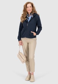 Felpa navy con chiusura a zip sul davanti, due tasche frontali e una texture morbida. Abbinata a pantaloni tapered beige chiaro e a una piccola borsa beige.