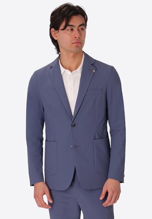 Uomo che indossa una giacca e pantaloni blu sopra una camicia bianca abbottonata, che guarda leggermente di lato su uno sfondo neutro.