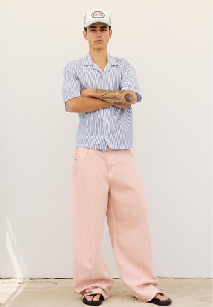 Jeune homme les bras croisés, portant une casquette blanche, une chemise rayée bleue à manches courtes, un pantalon large rose et des sandales noires, debout sur un sol en béton.
