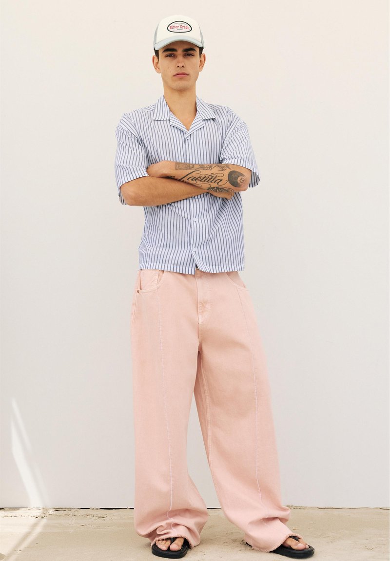 Jeune homme les bras croisés, portant une casquette blanche, une chemise rayée bleue à manches courtes, un pantalon large rose et des sandales noires, debout sur un sol en béton.