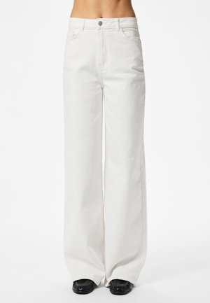 Persona que lleva puestos pantalones blancos de talle alto y pierna ancha con cierre de botón y cremallera, de pie con zapatos negros visibles bajo los pantalones.