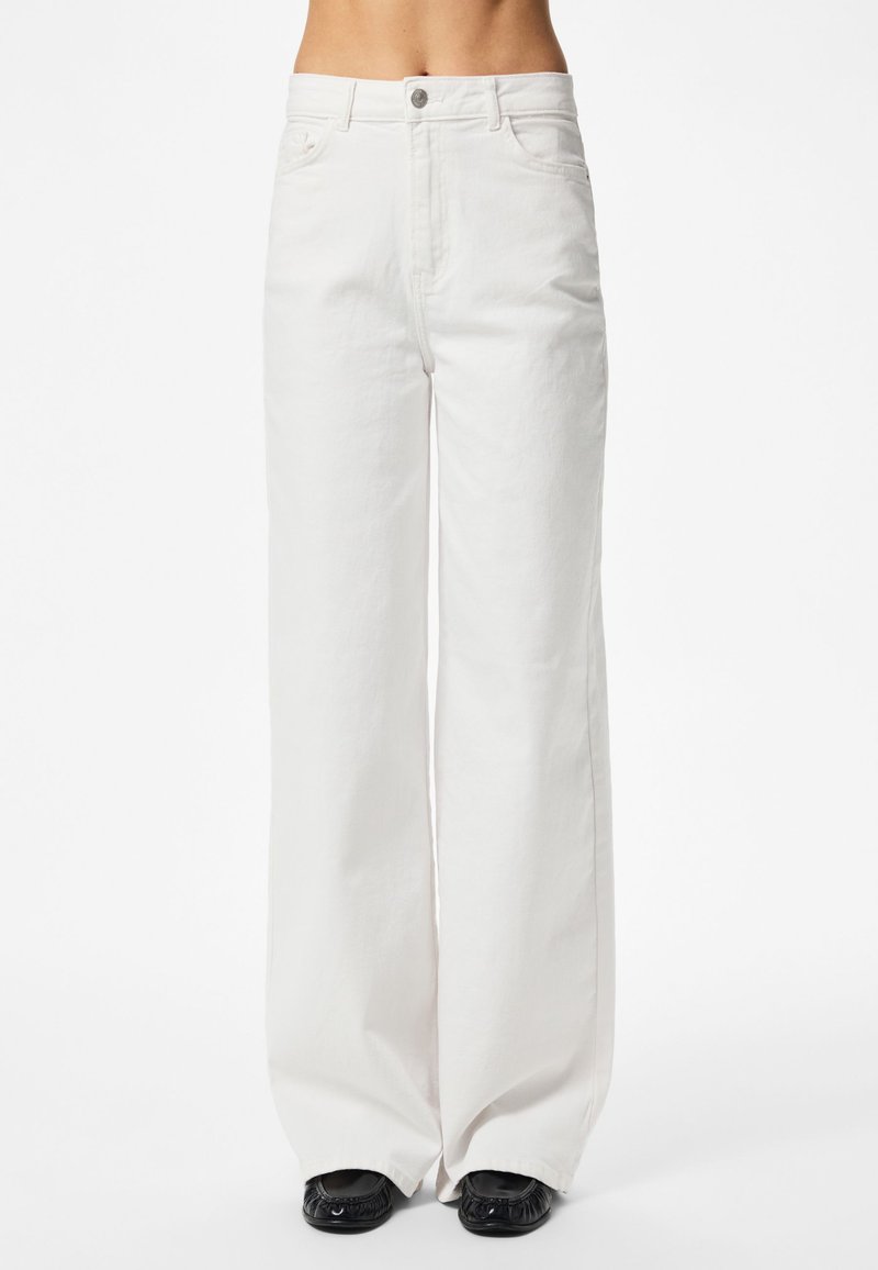 Personne portant un pantalon blanc taille haute à jambes larges, avec une fermeture par bouton et zip, debout avec des chaussures noires visibles sous le pantalon.