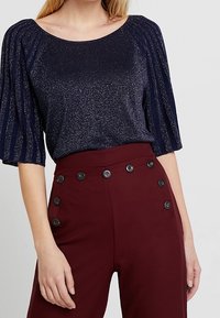 Marineblauer Glitzertop mit kurzen, plissierten Ärmeln, kombiniert mit hoch taillierten bordeauxroten Hosen, die schwarze Knopf-Details an der Taille aufweisen.