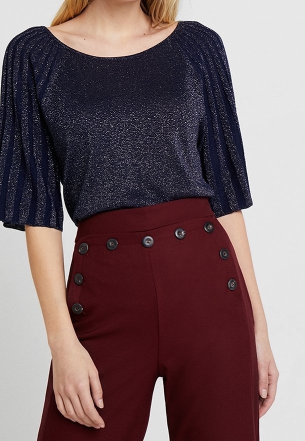 Marineblauer Glitzertop mit kurzen, plissierten Ärmeln, kombiniert mit hoch taillierten bordeauxroten Hosen, die schwarze Knopf-Details an der Taille aufweisen.