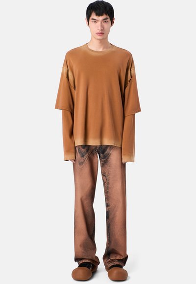 Lockerer brauner Pullover mit weicher Textur und braunen Jeans mit einem dunklen abstrakten Muster. Abgerundete braune Schuhe runden das Outfit ab.
