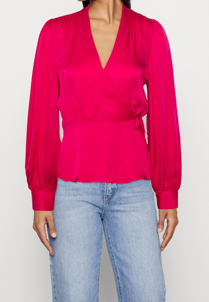 Jasně růžová zavinovací halenka z lesklé látky s dlouhými nadýchanými rukávy a zavazováním v pase. Má hluboký výstřih do V a hladkou, měkkou texturu.
