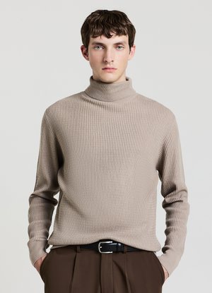 Giovane uomo con capelli corti castani che indossa un maglione a collo alto beige a coste e pantaloni marroni, in piedi con le mani in tasca.