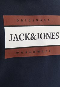 Tejido azul marino con un logotipo rectangular en blanco y rojo óxido. El texto dice "JACK&JONES" en negro, con "ORIGINALS" y "WORLDWIDE" debajo.