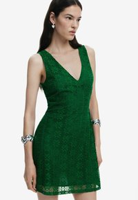 Grünes Spitzenkleid mit V-Ausschnitt, taillierter Form, floralem Muster und einem Saum, der knapp über dem Knie endet. Silberne Armkette als Akzent.