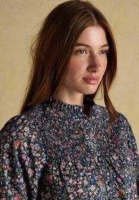 Jauna sieviete ar gariem brūniem matiem, ģērbusies augsta kakla ziedu blūzē ar piegriezuma detaļām, skatās nedaudz sānis pret vienmērīgu fona.