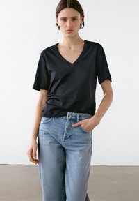 Kvinde iført en sort V-hals T-shirt og lyseblå jeans, stående indendørs med den ene hånd i lommen og et neutralt udtryk.