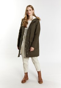 Oliven grøn parka med faux fur-krave, lynlås og trykknaplukning, sidelommer og en afslappet pasform, parret med cremede bukser og brune støvler.