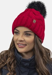 Red knitted beanie hat with a textured pattern, featuring a black faux fur pom-pom on top and a small fabric label on the side.