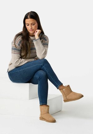 Stivali alla caviglia in suede beige con interno morbido, indossati con jeans blu e un maglione grigio chiaro con un design a motivo sulla parte superiore.