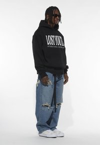 Black hoodie with white text, paired with distressed blue jeans featuring large rips. White sneakers complete the outfit.