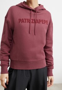 Felpa corta bordeaux con cappuccio e cordini, dotata di logo impresso "PATRIZIA PEPE" sul davanti. Tessuto morbido e polsini e orlo a coste. Visibile un pantalone nero.