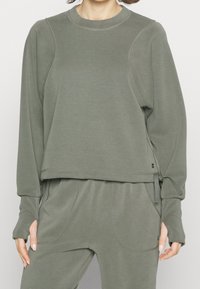 Sudadera verde caqui con mangas largas, escote redondo y aberturas laterales. Confeccionada con tela suave y detalles de costuras visibles. Combinada con pantalones a juego.