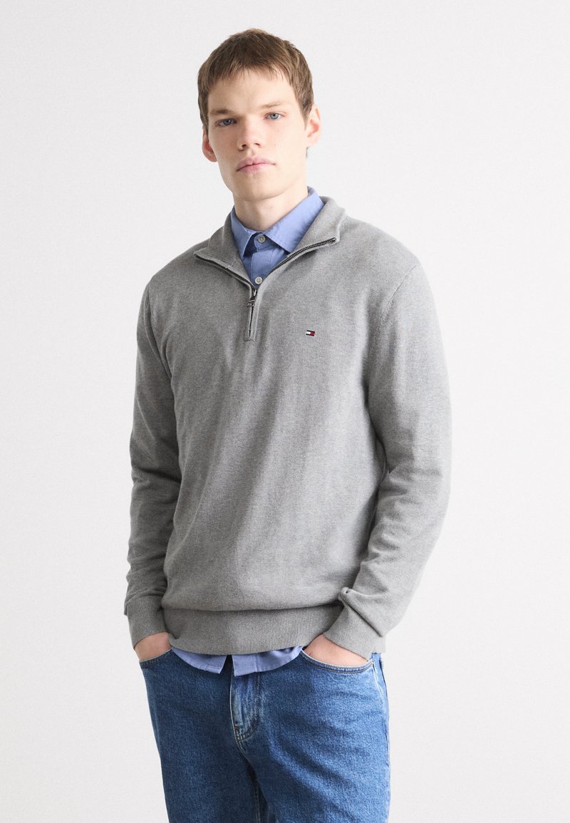 Pull gris zippé en matière tricot douce, avec un col contrastant, des poignets côtelés et un petit logo sur la poitrine.