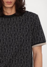 Svart T-skjorte med teksturert, repetitivt bokstavmønster. Korte ermer med ribbede, beige detaljer ved mansjettene og i halsen.