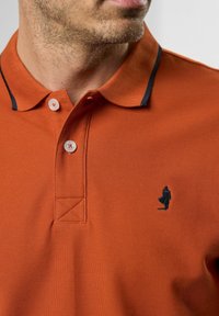 Narančasta polo majica od teksturirane tkanine s crnim ovratnikom i detaljima. Ima dva bijela gumba i mali crni logo na prsima.