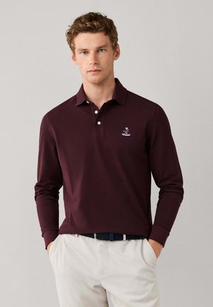 Joven con polo de manga larga color burdeos oscuro y pantalones beige claros con las manos en los bolsillos frente a un fondo liso.