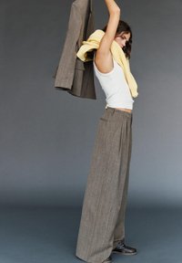 Kvinde iført brede grå bukser og hvid ærmeløs top løfter matchende jakke over hovedet, gul sweater hængende rundt om halsen, grå baggrund.