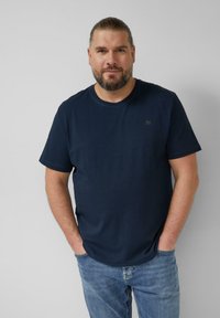 Homme avec barbe et cheveux attachés, portant un t-shirt bleu marine et un jean bleu, debout les mains dans les poches sur un fond uni.